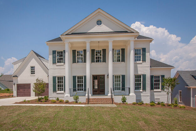 Large two-story white brick new construction home with four tall columns, black shutters, multiple windows, and a front porch. A manicured lawn and driveway are visible in front of the home, with clear skies overhead. This impressive property showcases the quality craftsmanship of Midlands contractors who specialize in building stunning new homes.