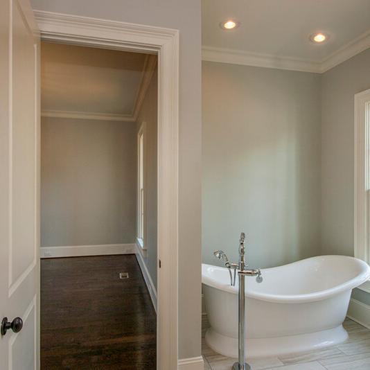 A bright bathroom with a white freestanding tub, gray walls, and a large window; an open door leads to an empty room with dark wood floors—an inviting space that perfectly exemplifies the quality and style found in new construction homes by Midlands contractors.