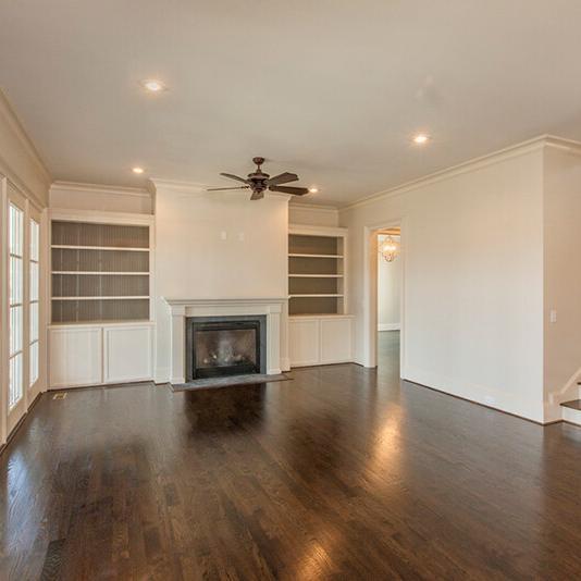 Bright, empty living room with dark wood floors, white walls, built-in shelves, a fireplace, ceiling fan, large windows, and stairs leading upwards—an inviting space that showcases the high-quality craftsmanship of Midlands contractors in this beautiful new construction home.