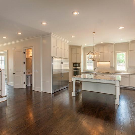 Spacious kitchen with white cabinets, stainless steel appliances, a center island, pendant lights, and dark wood floors. Perfectly designed by custom home builders, this new construction home features a staircase on the left and large windows that let in an abundance of natural light.