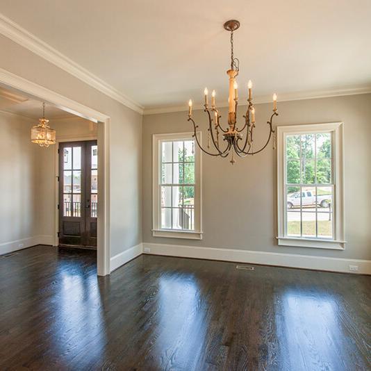 Empty dining room with dark hardwood floors, beige walls, large windows letting in natural light, and a chandelier hanging from the ceiling. Featuring a custom floor plan by Midlands contractors, this inviting space seamlessly connects to an adjacent room with a pendant light visible through a wide doorway.