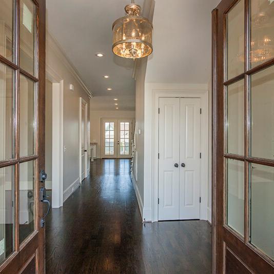 View through open wooden French doors into a hallway with dark wood floors, white walls, a modern light fixture, and doors leading to other rooms; windows are visible at the end of the hall—a stunning example of craftsmanship by custom home builders in this new construction home.
