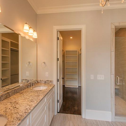 A modern bathroom in a new construction home featuring a granite double sink, large mirror, glass shower, and stylish light fixtures above the sinks. An open doorway leads to a spacious walk-in closet with shelving—ideal for those seeking the thoughtful design offered by custom home builders.