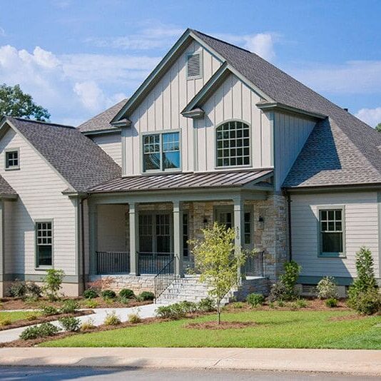 Two-story gray new construction home with a gabled roof, large front porch, columns, stone accents, and well-maintained landscaping, set against a backdrop of trees and a blue sky. This beautifully designed residence offers the opportunity for a custom floor plan to perfectly suit your lifestyle.