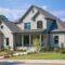 Two-story gray new construction home with a gabled roof, large front porch, columns, stone accents, and well-maintained landscaping, set against a backdrop of trees and a blue sky. This beautifully designed residence offers the opportunity for a custom floor plan to perfectly suit your lifestyle.