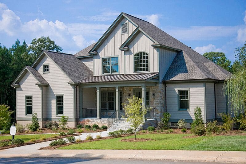 Two-story gray new construction home with a gabled roof, large front porch, columns, stone accents, and well-maintained landscaping, set against a backdrop of trees and a blue sky. This beautifully designed residence offers the opportunity for a custom floor plan to perfectly suit your lifestyle.