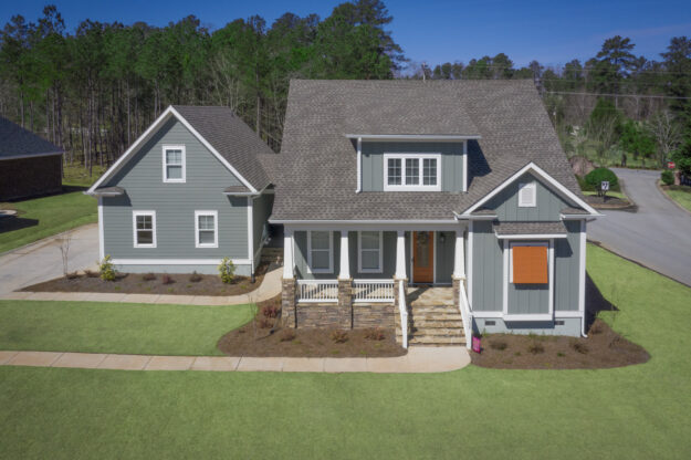 A gray two-story new construction home with white trim, a covered porch, and an attached garage, surrounded by neatly trimmed grass and landscaping, set in a suburban neighborhood near a wooded area.
