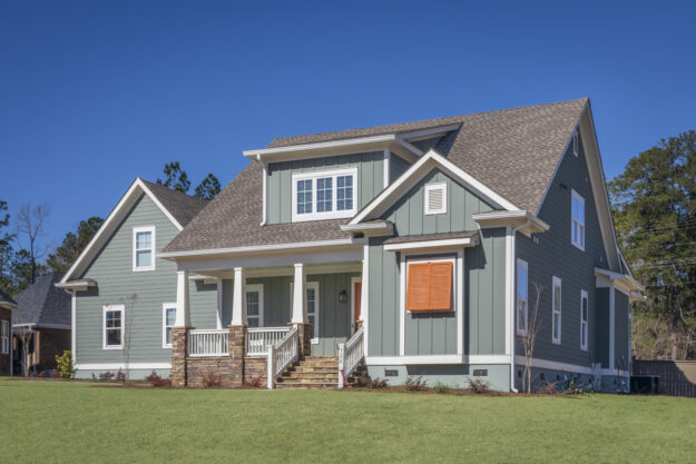 A modern two-story new construction home with green siding, white trim, a covered front porch, and wooden steps, set on a grassy lawn under a clear blue sky. Crafted by Midlands contractors, this residence features a custom floor plan designed for comfortable living.