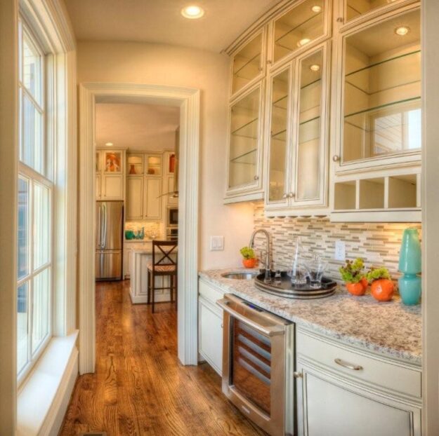 Bright, narrow kitchen with glass-front cabinets, granite countertops, a wine fridge, decorative bowls, and a sink. Hardwood floors—part of the custom floor plan by Midlands contractors—lead to a larger kitchen area with barstools in the background of this beautiful new construction home.