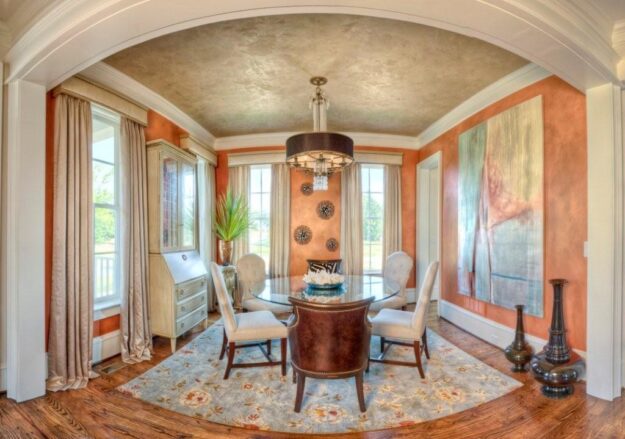A dining room with a round glass table, five chairs, orange walls, a large abstract painting, a chandelier, tall windows, and decorative vases on a patterned rug—perfect for those seeking inspiration for their new construction home.