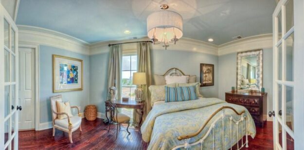 A cozy bedroom in a new construction home features a vintage bed, pastel bedding, wooden floor, dresser with mirror, window with drapes, armchair, artwork on the wall, and a chandelier hanging from the ceiling.
