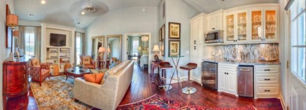 A bright, elegant living area designed by custom home builders features a beige sofa, armchairs, TV, built-in shelves, a small bar with glass cabinets, and large windows; hardwood floors and rugs accent the space.