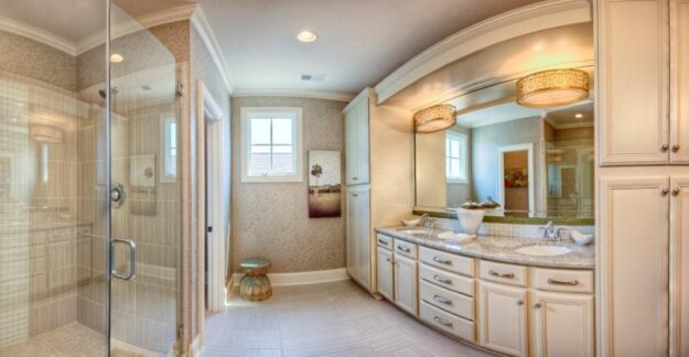 Elegant bathroom with glass shower, double sinks, large mirror, cream cabinets, two pendant lights, a window, artwork, and a small round side table. Neutral tones and ample lighting create a bright, spacious feel—perfect inspiration for new homes or anyone exploring a custom floor plan to achieve timeless style and comfort.