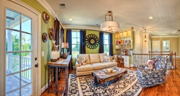 A cozy living room in this new construction home features a beige sofa, patterned armchair, wooden coffee table, decorative rug, wall art, and shelves; large windows let in natural light, and a French door opens to a porch.