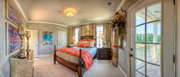 A bright, elegant bedroom in a new construction home features a large wooden bed, colorful bedding, artwork on the walls, tall candles, and glass doors opening to a sunny balcony with a view of greenery outside.
