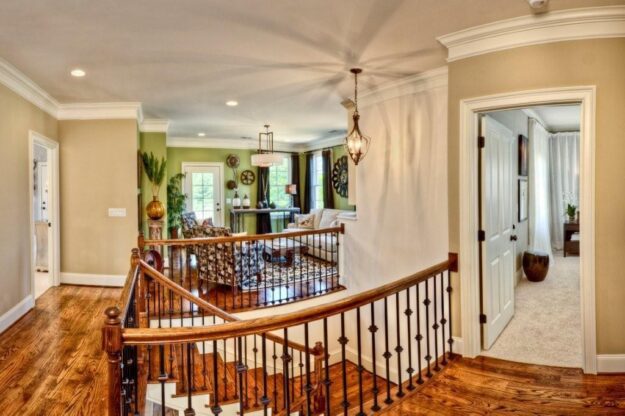 A bright hallway with wooden floors and an iron-railed staircase leads to a cozy living area with large windows, armchairs, and plants in this beautifully designed new construction home. A doorway on the right opens to a softly lit bedroom with light curtains, showcasing the thoughtful custom floor plan created by Midlands contractors.