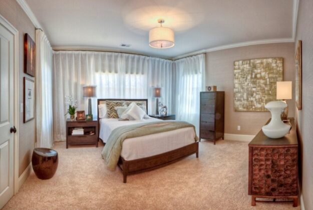 A modern bedroom with a large bed, nightstands, lamps, dresser, abstract art, and sheer curtains covering two walls—crafted by Midlands contractors as part of stunning new homes. Soft beige carpet and warm lighting complete this inviting space, offering both style and comfort with a custom floor plan designed to suit your lifestyle.