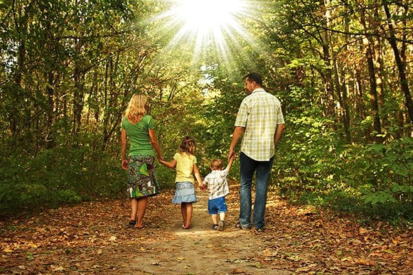 A family of four walks hand in hand down a forest path, sunlight streaming through the trees ahead—a perfect moment that feels right at home in one of our beautiful new construction homes.
