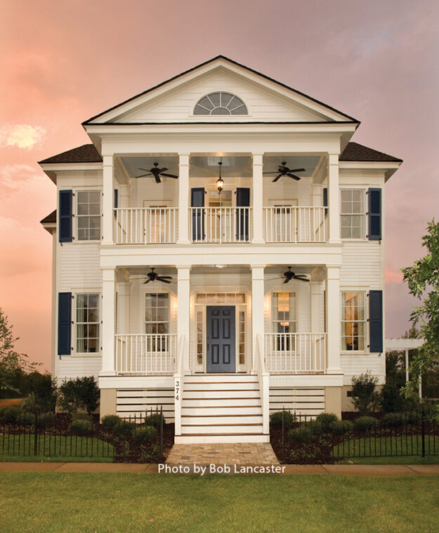 A two-story white house with blue shutters, double front porches, black ceiling fans, and a central front staircase—crafted by Midlands contractors—stands against a sunset sky, offering the charm and comfort of a new home with a custom floor plan tailored to your lifestyle.