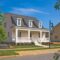 White two-story house with dormer windows and a covered front porch, set on a grassy lawn with a concrete walkway. Featuring a custom floor plan crafted by reputable Midlands contractors, this inviting home has a tree in the foreground and another house visible to the right.