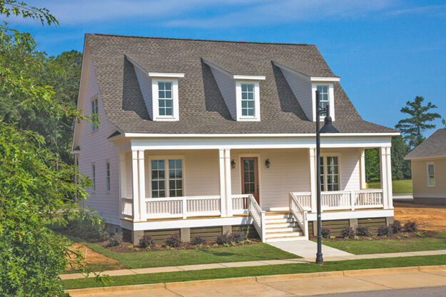A two-story white house with a large front porch, three dormer windows on the roof, and a small front yard with grass and shrubs, set on a corner lot under a clear blue sky, showcases the charm of new construction homes built by Midlands contractors.