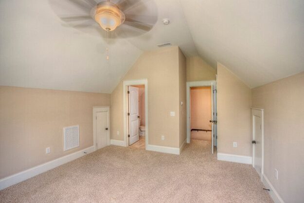 A carpeted attic room with beige walls, a ceiling fan, two small doors, and two open doorways—one leading to a bathroom and the other to a hallway. The room features sloped ceilings and white trim, showcasing the thoughtful design often found in new construction homes crafted by skilled custom home builders.