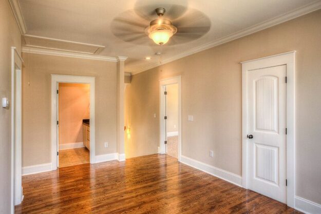 Empty room with beige walls, wooden floor, ceiling fan with light, and three white doors. One door leads to a bathroom, and another opens to a carpeted room. Crown molding decorates the ceiling—an inviting blank slate ideal for those seeking a custom floor plan from experienced custom home builders who specialize in beautiful new homes.