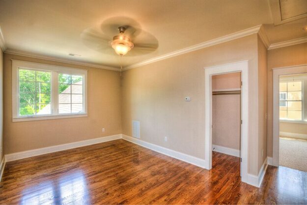Bright empty room with beige walls, wood floor, white trim, ceiling fan with light, two large windows, and an open closet door. Sunlight streams in, creating a warm, inviting atmosphere—an ideal space in a new construction home ready for your custom floor plan.
