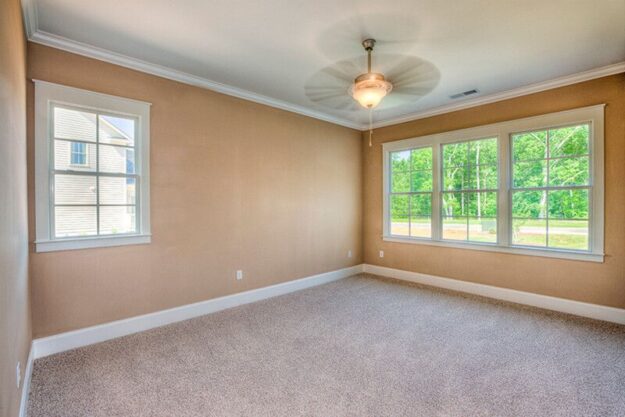 Empty beige room with a carpeted floor, large windows on one wall, a smaller window on another wall, white trim, and a ceiling fan with a light. Lush greenery is visible outside the windows in this new construction home—expertly crafted by Midlands contractors—with a custom floor plan designed for modern living.