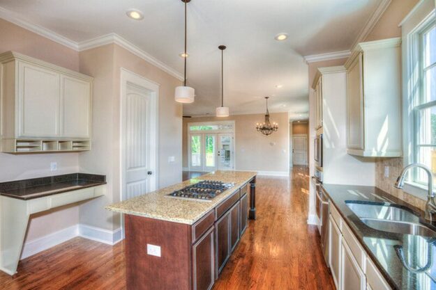 Bright kitchen with granite island, gas cooktop, white cabinets, desk nook, stainless sink, hardwood floors, hanging lights, and large windows. Designed as part of a custom floor plan by leading custom home builders, this space showcases thoughtful details like a chandelier and glass doors visible in the adjoining room.