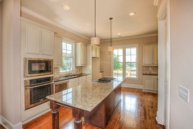Bright kitchen with beige cabinets, stainless steel appliances, granite island with built-in stovetop, pendant lights, and large windows letting in natural light. Hardwood floors add warmth to the contemporary space—an inviting centerpiece often found in new homes crafted by leading Midlands contractors.
