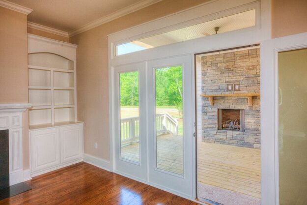 A bright room with wooden floors, built-in white shelves, and large glass doors that open to a patio with a stone fireplace and green trees outside—an inviting space often found in new homes designed by custom home builders.