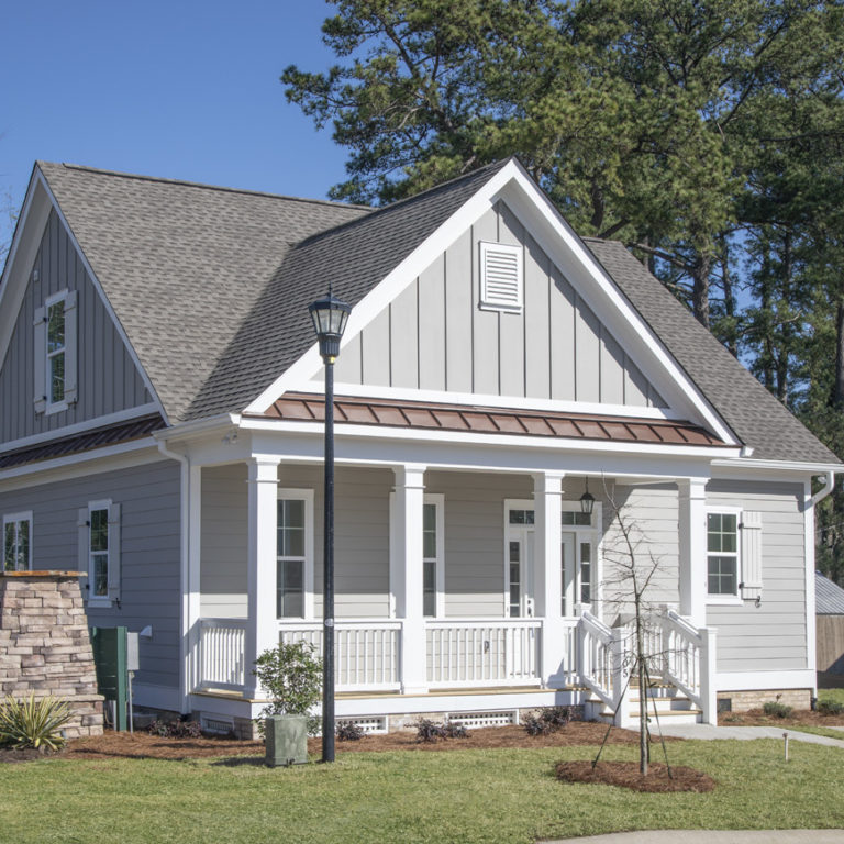 WEB_02_1105_Congaree_Bluff_73A3953 A small, modern gray house with a porch, white trim, gabled roof, and chimney—crafted by experienced custom home builders—set in a green yard with trees and landscaping under a clear blue sky.