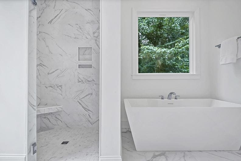 Modern bathroom with a white freestanding tub, a large window showing green trees, and a marble-tiled walk-in shower with a built-in shelf and bench. Designed by Midlands contractors, this space showcases the thoughtful touches found in new homes with custom floor plans. Towels hang on the right wall.
