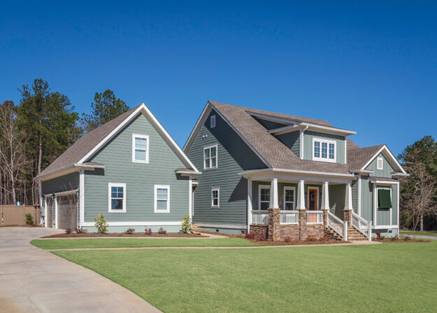 A large, modern gray house with white trim and a detached garage, set on a manicured lawn under a clear blue sky, showcases the craftsmanship of custom home builders, with trees in the background.