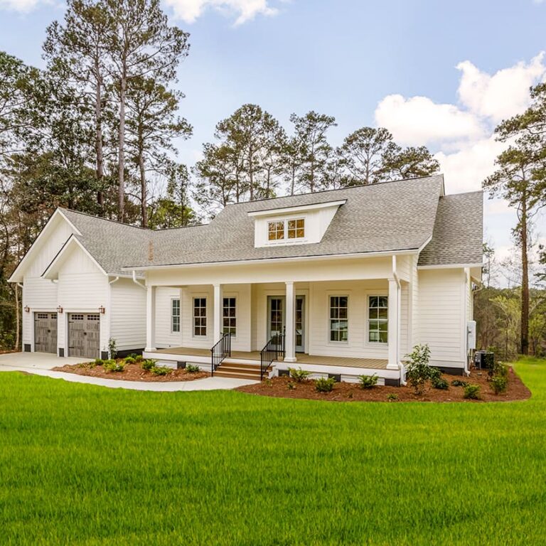 New-Homes-In-Arcadia-Bluff-Lady-Street-Builders A white, modern farmhouse with a front porch, attached garage, and large windows sits on a green lawn, surrounded by tall trees under a partly cloudy sky—an inviting new construction home expertly crafted by custom home builders in partnership with Midlands Contractors.