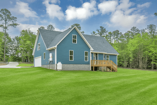 A blue, two-story house with a gray roof, front porch, and attached garage sits on a large, well-manicured lawn surrounded by trees under a partly cloudy sky—an impressive example of what midlands contractors can achieve when working with a custom floor plan tailored to your needs.