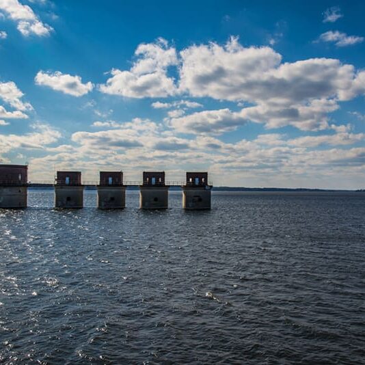 Five square concrete structures stand in a row over a large body of water under a partly cloudy sky, with sunlight reflecting on the rippling surface—a striking reminder of the innovation and strength that custom home builders bring to each new construction home.