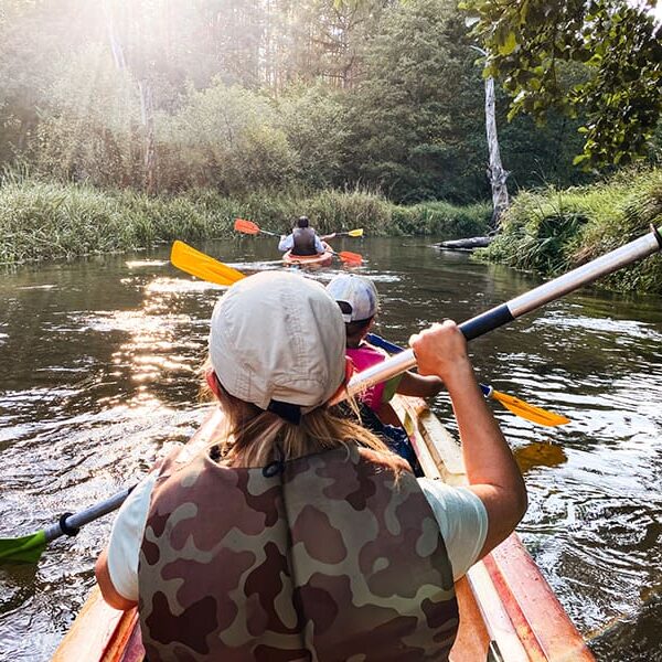 Rivers-Of-Cola-Lady-Street-Builders A group of people kayaking down a calm river surrounded by green trees and grass, with sunlight shining through the foliage—much like exploring new possibilities with custom home builders designing your perfect retreat.