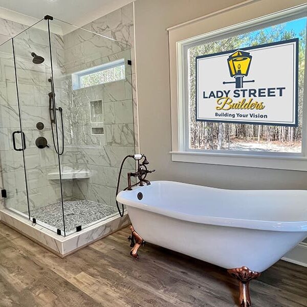 Top-Custom-Home-Features-Lady-Street-Builders Modern bathroom with a glass shower, white freestanding tub with bronze feet, wood flooring, and a large window displaying the Lady Street Builders logo and slogan—perfectly showcasing the attention to detail you can expect from custom home builders. This inviting space highlights the possibilities available in your new construction home or custom floor plan.