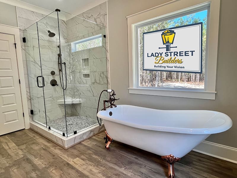 Top-Custom-Home-Features-Lady-Street-Builders Modern bathroom with a glass shower, white freestanding tub with bronze feet, wood flooring, and a large window displaying the Lady Street Builders logo and slogan—perfectly showcasing the attention to detail you can expect from custom home builders. This inviting space highlights the possibilities available in your new construction home or custom floor plan.