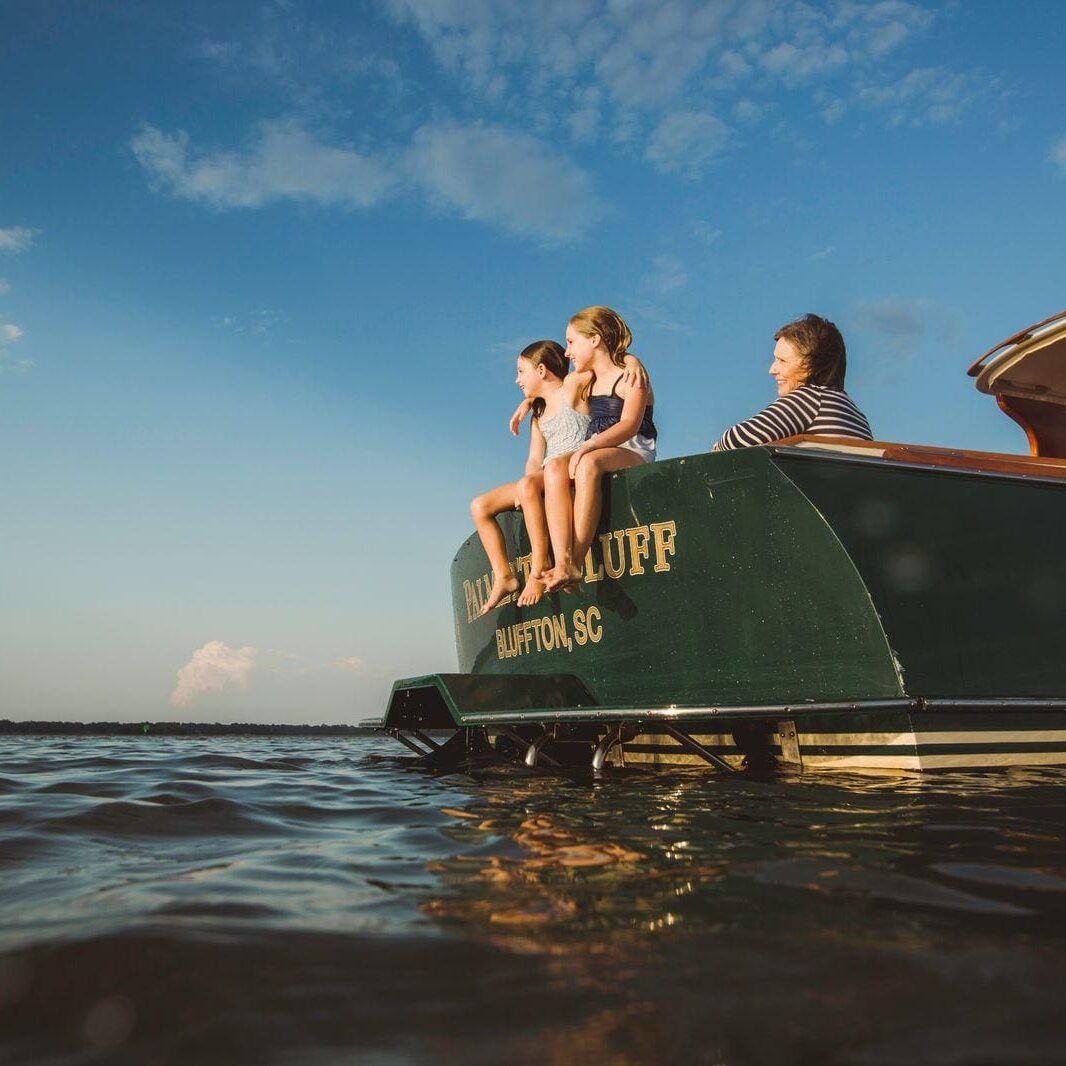 Lake Murray Living Lady Street Builders Three people sit on the back of a green boat named Bluffton SC enjoying the sunny weather, with their feet dangling above the water and a calm, blue sky in the background—perhaps taking a break from exploring new homes or dreaming up ideas for their own new construction home with Midlands Contractors.