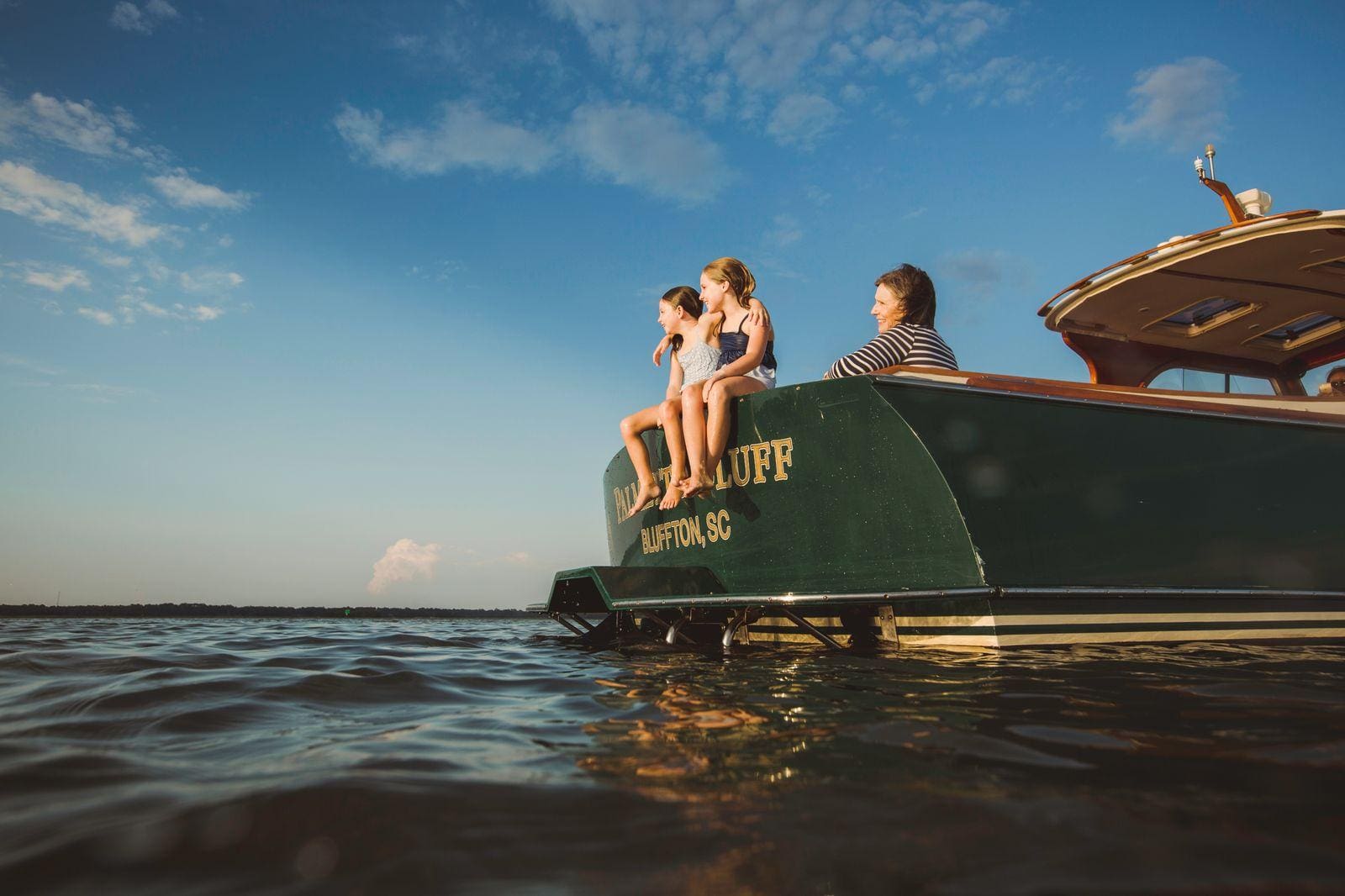 Lake Murray Living Lady Street Builders Three people sit on the back of a green boat named Bluffton SC enjoying the sunny weather, with their feet dangling above the water and a calm, blue sky in the background—perhaps taking a break from exploring new homes or dreaming up ideas for their own new construction home with Midlands Contractors.