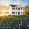 A two-story white new construction home with columns stands by a lake at sunset. Overlaid in the grass is a blueprint floor plan created by Midlands contractors, showing labeled rooms like great room, kitchen, and primary suite.