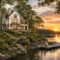 A cozy lakeside house with a custom floor plan and large windows sits among lush greenery at sunset, overlooking a calm lake. A wooden dock with chairs and a small boat extends into the water, reflecting the golden sky.