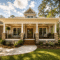 A charming house with a wide front porch, white columns, stone accents, and a wooden door—crafted by skilled Midlands contractors—sits surrounded by lush lawns, vibrant plants, and tall trees under a blue sky with scattered clouds. This inviting property reflects the quality and expertise of custom home builders dedicated to creating beautiful new homes in the heart of the Midlands.