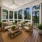 A cozy screened-in porch, crafted by experienced midlands contractors, features wicker furniture with beige cushions, green pillows, potted plants, a wooden coffee table, and a woven rug. Large windows reveal a lush, green outdoor view—perfect for those seeking custom home builders to create serene new construction home retreats.