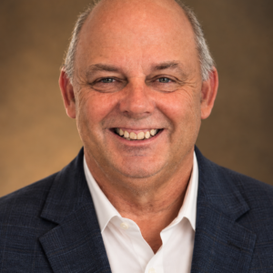Smiling local custom builder and contractor, Tony Carnaggio, poses wearing a white shirt and dark blue blazer, poses in front of a brown blurred background, representing the trusted expertise of Lady Street Builders, custom builder in Columbia.