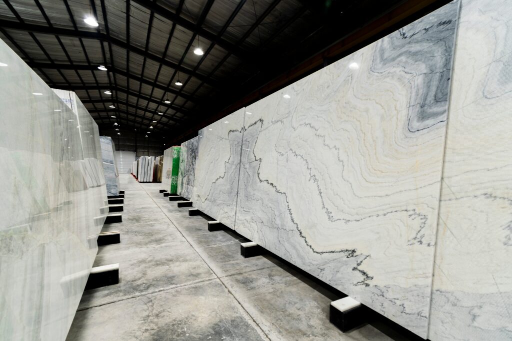 Large marble slabs with gray and white veining are displayed upright in rows inside a spacious, industrial warehouse with a high ceiling and overhead lighting, alongside stunning quartz countertops. Discover care tips and maintenance guidelines to ensure your surfaces remain beautiful for years to come.