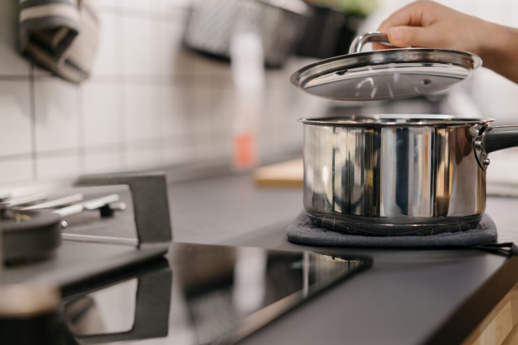 A hand lifts the lid off a stainless steel pot on a stovetop in a modern kitchen with quartz countertops, a tiled backsplash, and blurred kitchen items in the background.